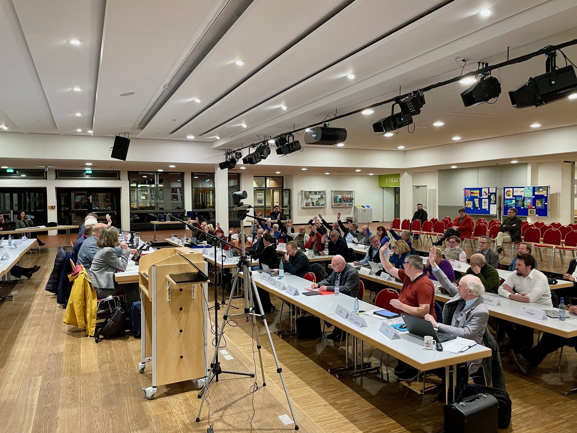 Viele Menschen sitzen in aufgereihten Tischen, schauen nach vorne und haben zur Abstimmung eine Hand gehoben.