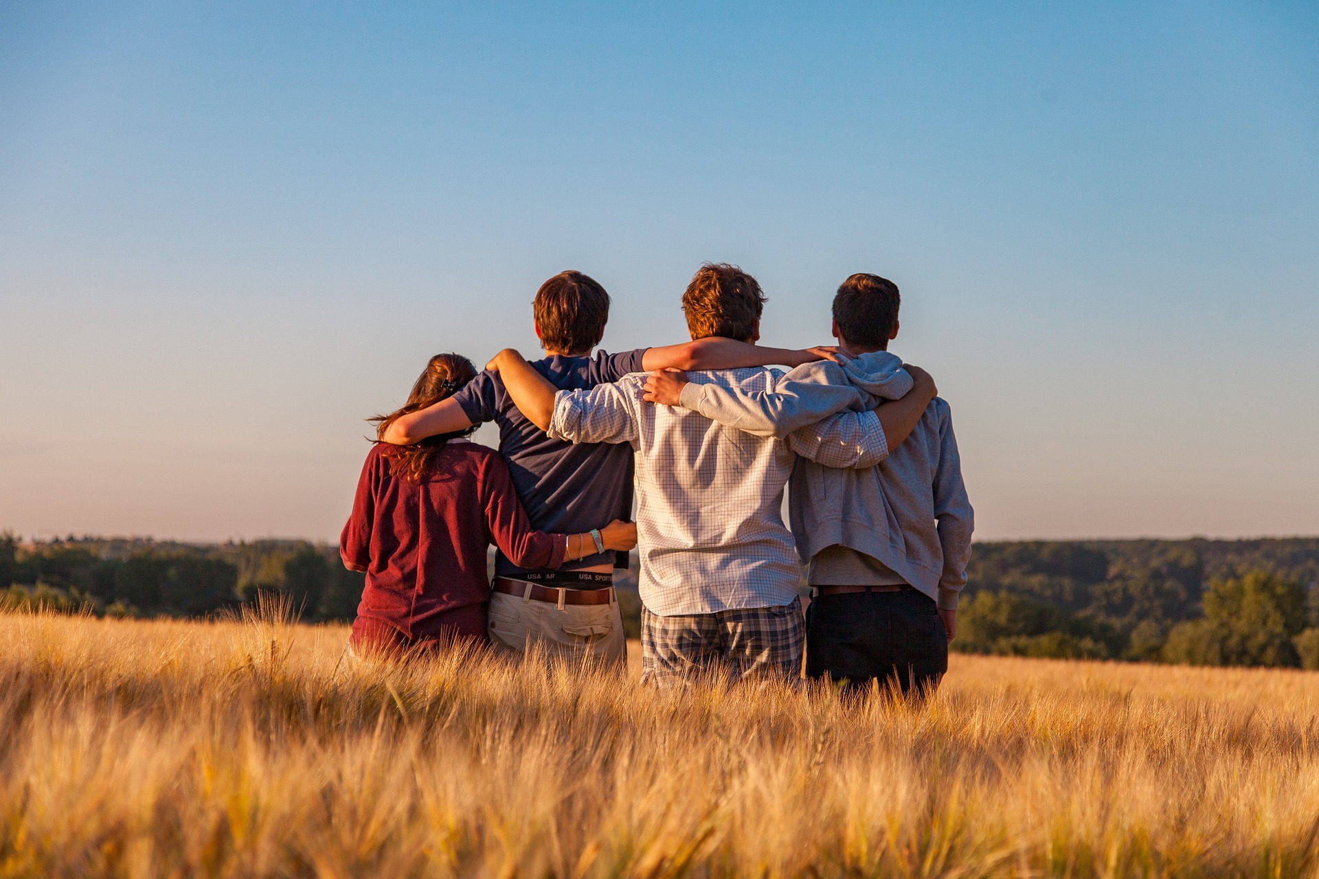 Vier Jugendlichen von hinten fotografiert, sitzen arm in arm auf einem Feld