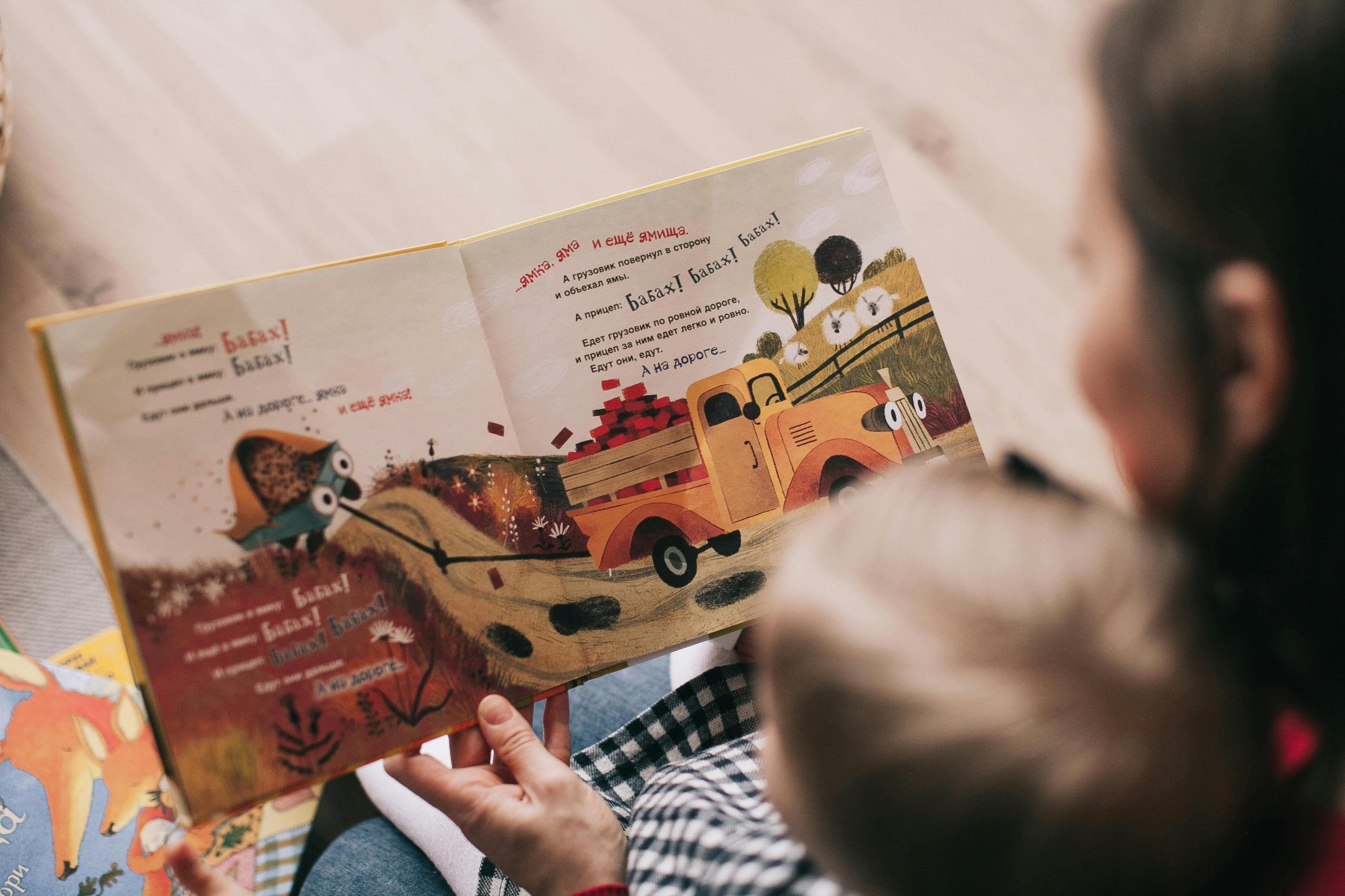 Eine Frau sitzt mit einem kleinen Kind vor einem Buch. Beide sind von hinten zu sehen.