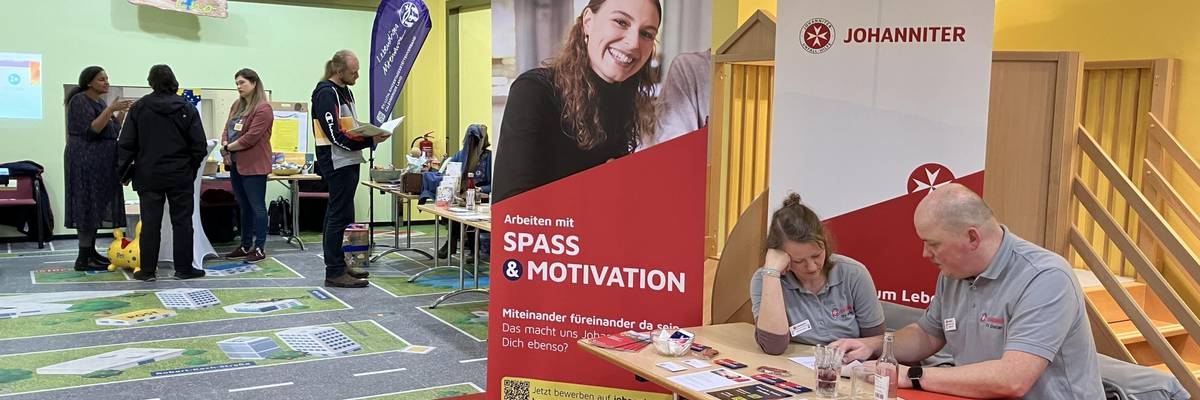 In der rechten Bildhälfte ist der Messestand der Johanniter mit 2 Personen zu sehen, die am Tisch sitzen und zu dem Stand gehören. An dem Stand stehen zwei Roll-ups der Johanniter. In der linken Bildhälfte sind im Hintergrund Aussteller und Besuchende zu sehen, die sich an den Messeständen informieren.