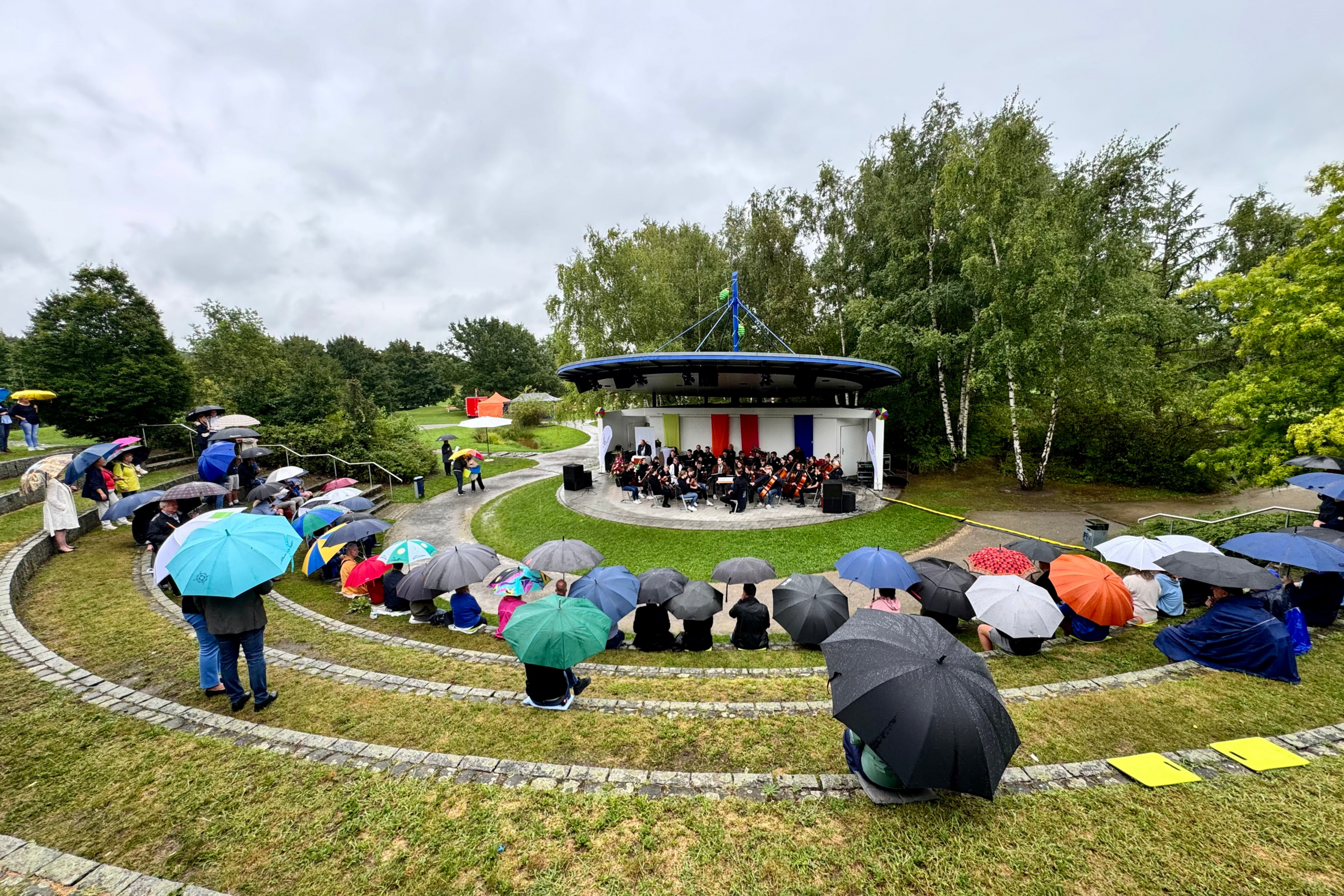 zahlreiche Menschen sitzen mit bunten Regenschirmen auf einer Steintreppe und blicken Richtung Bühne.