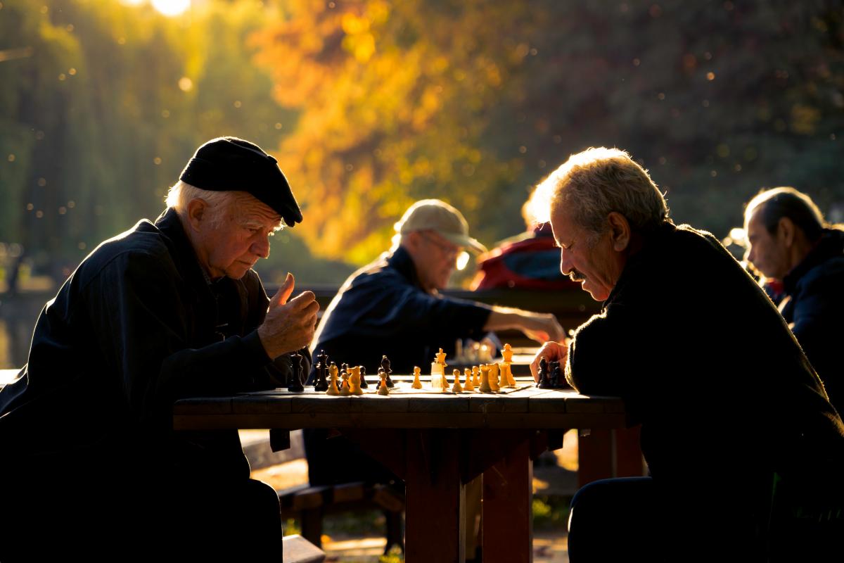 Männer sitzen an mehreren Tischen im Park und spielen Schach. Sie befinden sich in einem Park. Das Licht ist das einer sommerlichen Abenddämmerung.