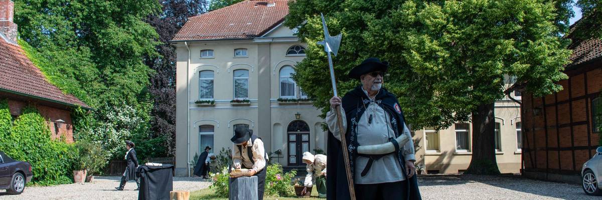 Ein verkleideter Mann winkt. Mehrere altertümlich gekleidete Personen befinden sich im Hintergrund.