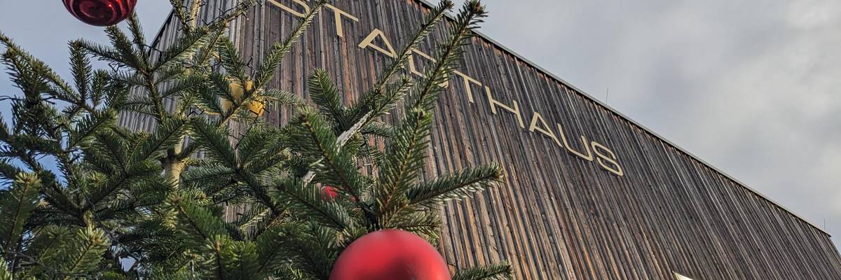 Ein grüner Weihnachtbaum mit roten und silbernen Kugeln geschmückt steht vor der Holzfassade des Stadthauses.