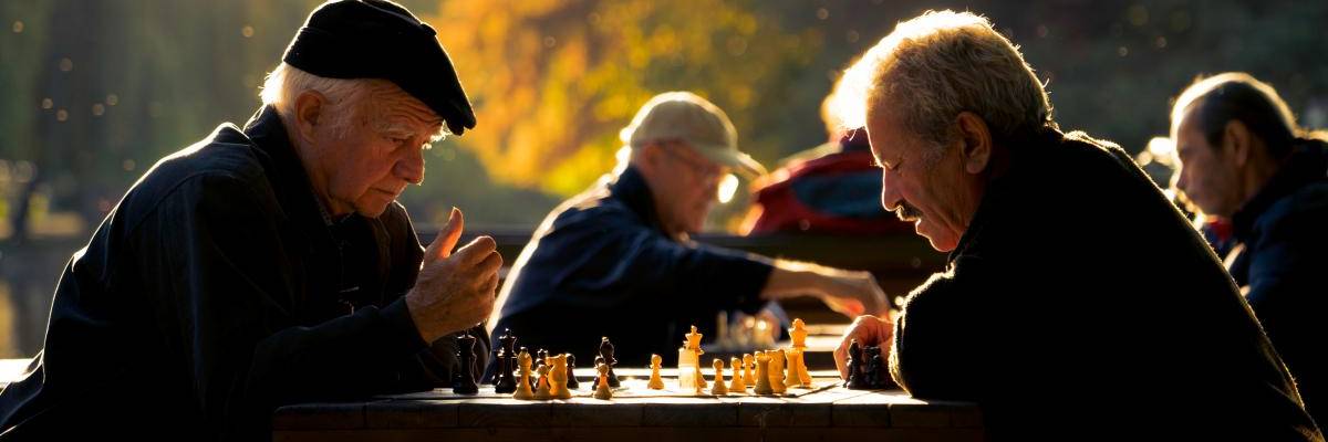 Männer sitzen an mehreren Tischen im Park und spielen Schach. Sie befinden sich in einem Park. Das Licht ist das einer sommerlichen Abenddämmerung.