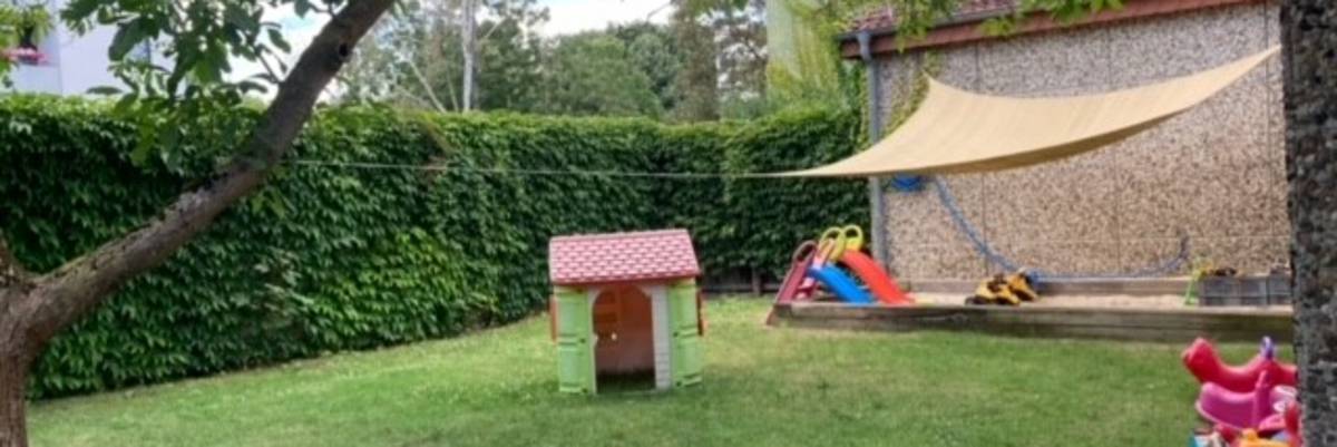 Garten der Großtagespflege Wichtelhöhle. Im hinteren Teil des Gartens steht ein Sandkasten, welcher mit einer Plane überspannt ist. Links steht ein Baum. In der Mitte des Gartens steht ein kleines Spielhäuschen. Rechts an der Hauswand stehen kleine Fahrzeuge für Kinder.