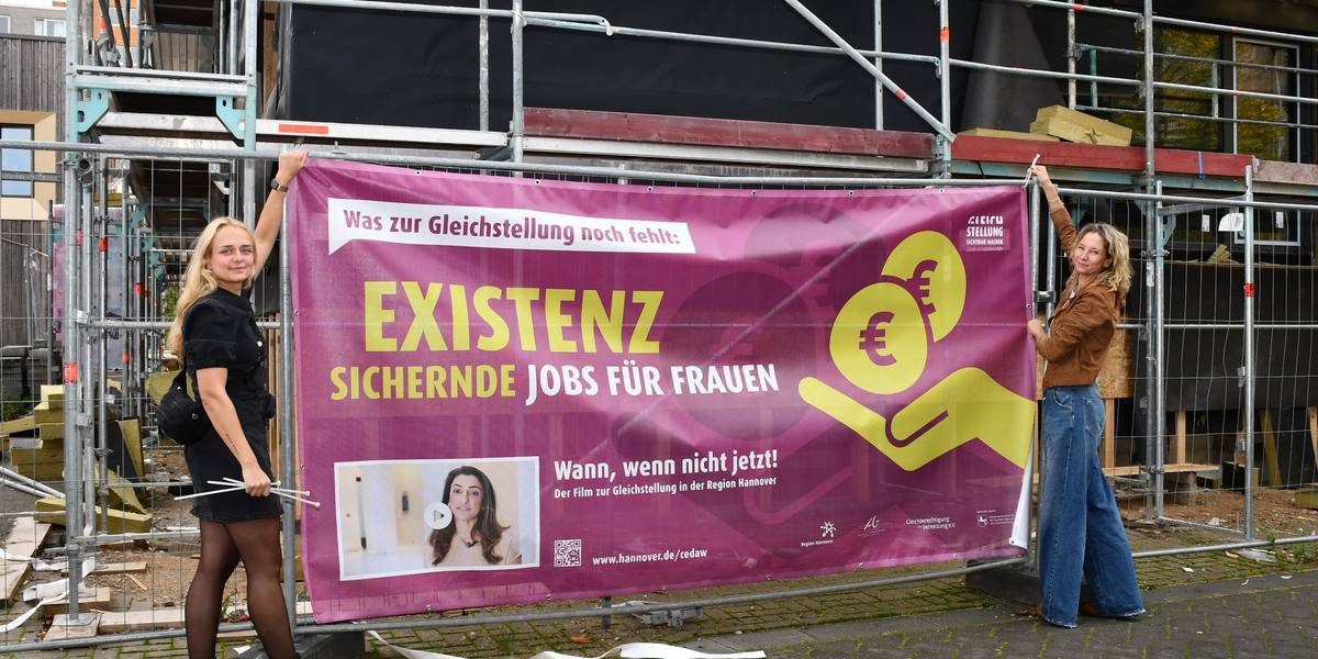 Zwei Frauen bringen ein Banner an einem Baustellenzaun an.