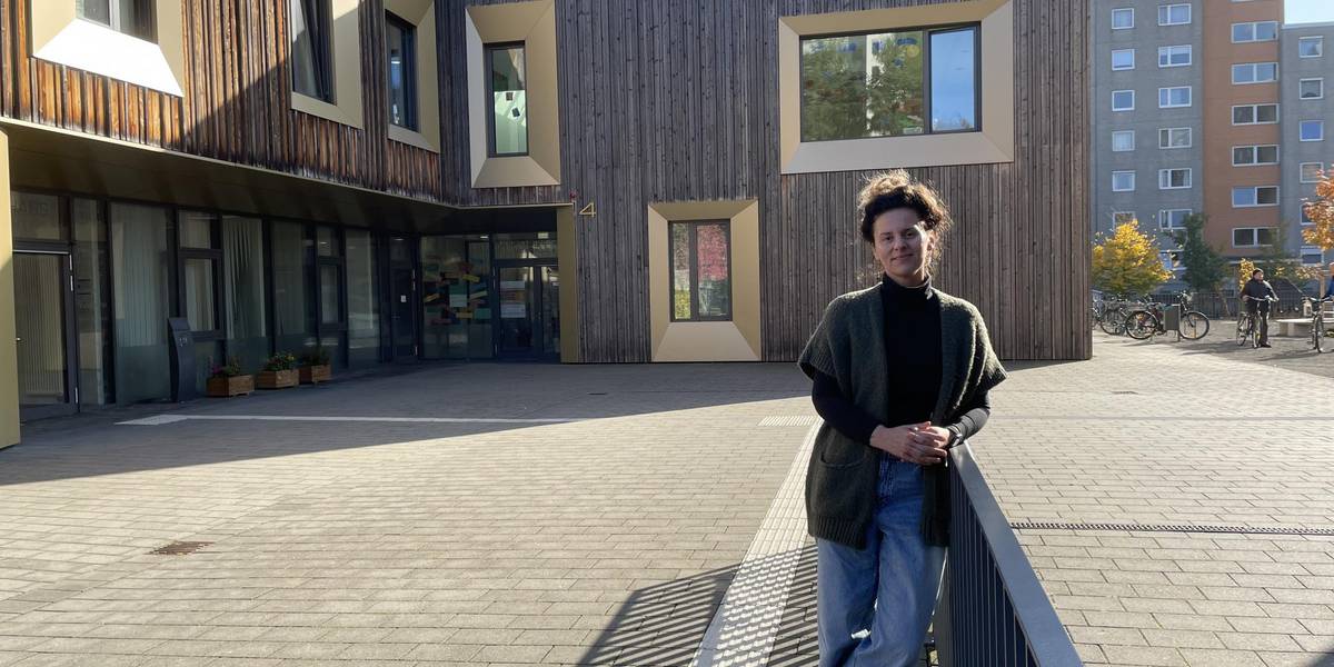 Eine Frau lehnt vor einem Gebäude mit holzverkleideter Fassade an einem Geländer und blickt freundlich in die Kamera.