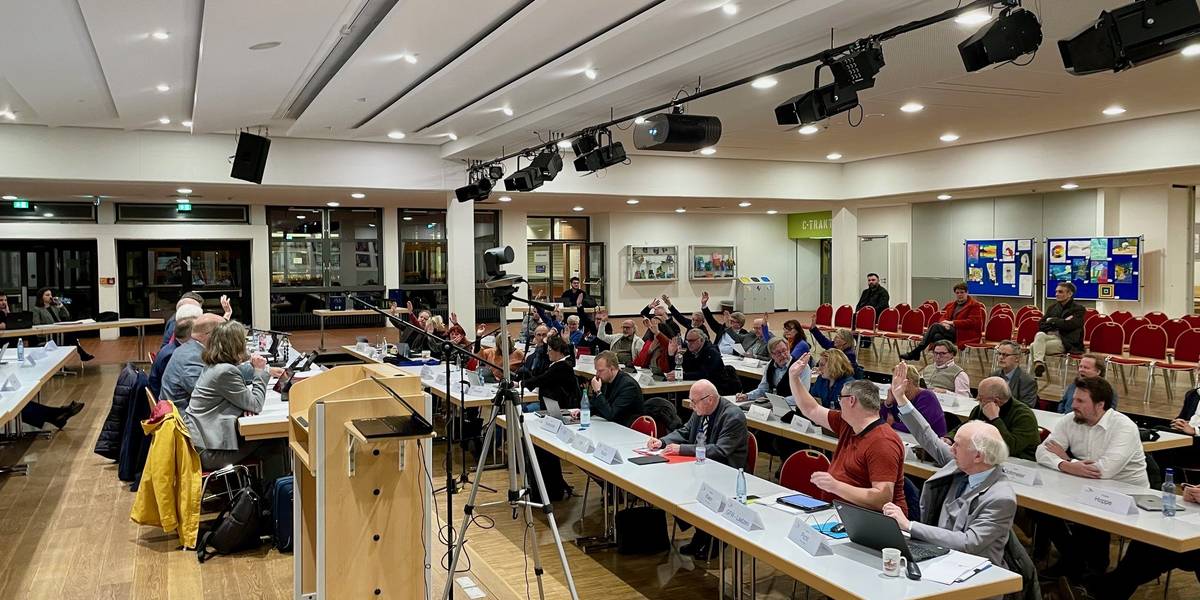 Viele Menschen sitzen in aufgereihten Tischen, schauen nach vorne und haben zur Abstimmung eine Hand gehoben.