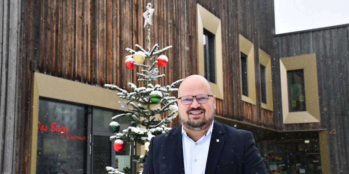 Ein Mann steht vor einem Holzhaus, vor dem ein geschmückter Tannenbaum steht. Es schneit.