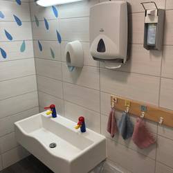 Im Badezimmer hängt auf Kinderhöhe ein Waschbecken mit bunten Wasserhähnen. Rechts neben dem Waschbecken hängt eine Leiste mit Haken für Waschlappen. Über dem Waschbecken hängt eine Lampe, ein Seifenspender, Desinfektionsspender und Papiertücher. Unter dem Waschbecken steht ein Mülleimer. In der linken Ecke an der Wand sind Wassertropfen aufgeklebt.