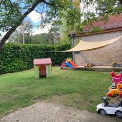 Im Garten steht ein Sandkasten, welcher mit einer Plane überspannt ist. Links steht ein Baum. Mitten auf der Wiese steht ein kleine Spielhäuschen. Rechts an der Hauswand stehen kleine Fahrzeuge für Kinder.