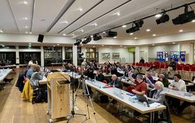 Viele Menschen sitzen in aufgereihten Tischen, schauen nach vorne und haben zur Abstimmung eine Hand gehoben.