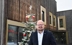 Ein Mann steht vor einem Holzhaus, vor dem ein geschmückter Tannenbaum steht. Es schneit.