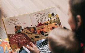 Eine Frau sitzt mit einem kleinen Kind vor einem Buch. Beide sind von hinten zu sehen.