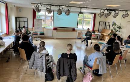 Die Teilnehmenden sitzen im Halbkreis auf stühlen und gucken zu einer Präsentationswand. Auf einer Leinwand wird eine Präsentation vorgestellt.