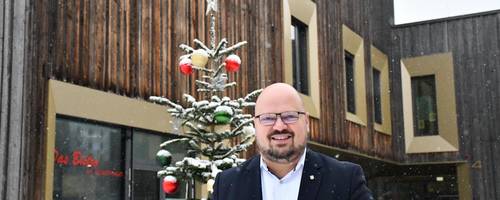 Ein Mann steht vor einem Holzhaus, vor dem ein geschmückter Tannenbaum steht. Es schneit.