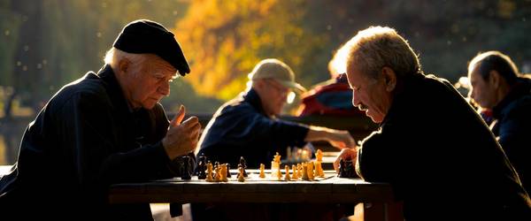 Männer sitzen an mehreren Tischen im Park und spielen Schach. Sie befinden sich in einem Park. Das Licht ist das einer sommerlichen Abenddämmerung.